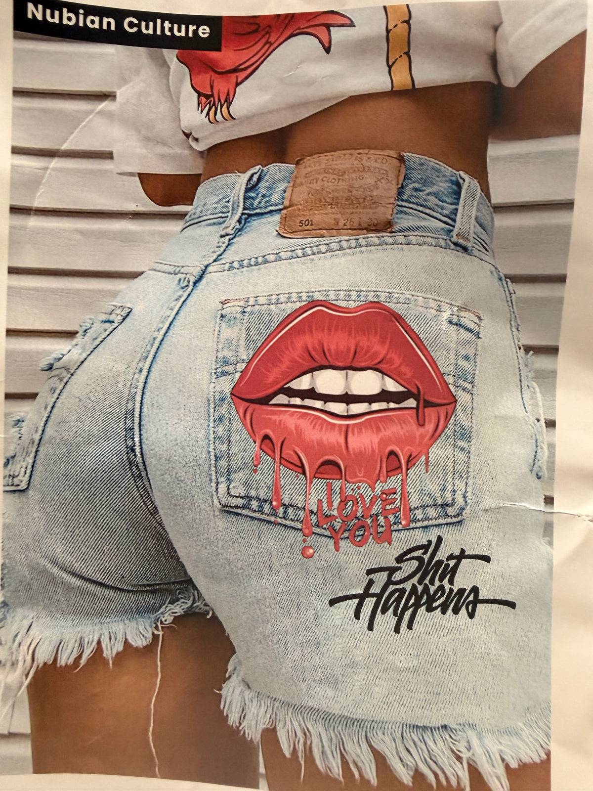 A close-up of a person wearing light-wash denim cutoff shorts with a large red lips graphic and dripping text on the back pocket, paired with a white graphic T-shirt. The photo is cropped tightly on the lower torso and upper thighs, with a sliver of a white slatted wall in the background.