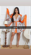 A woman modeling a short orange and white patterned kimono-style dress with wide sleeves, standing in a bright indoor room with a side table and chair visible.