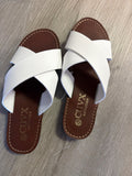 A pair of white leather slide sandals with brown footbeds, featuring crossed straps and the brand name 'CUVX collection' printed on the insole, laid flat on a gray wood-textured surface.