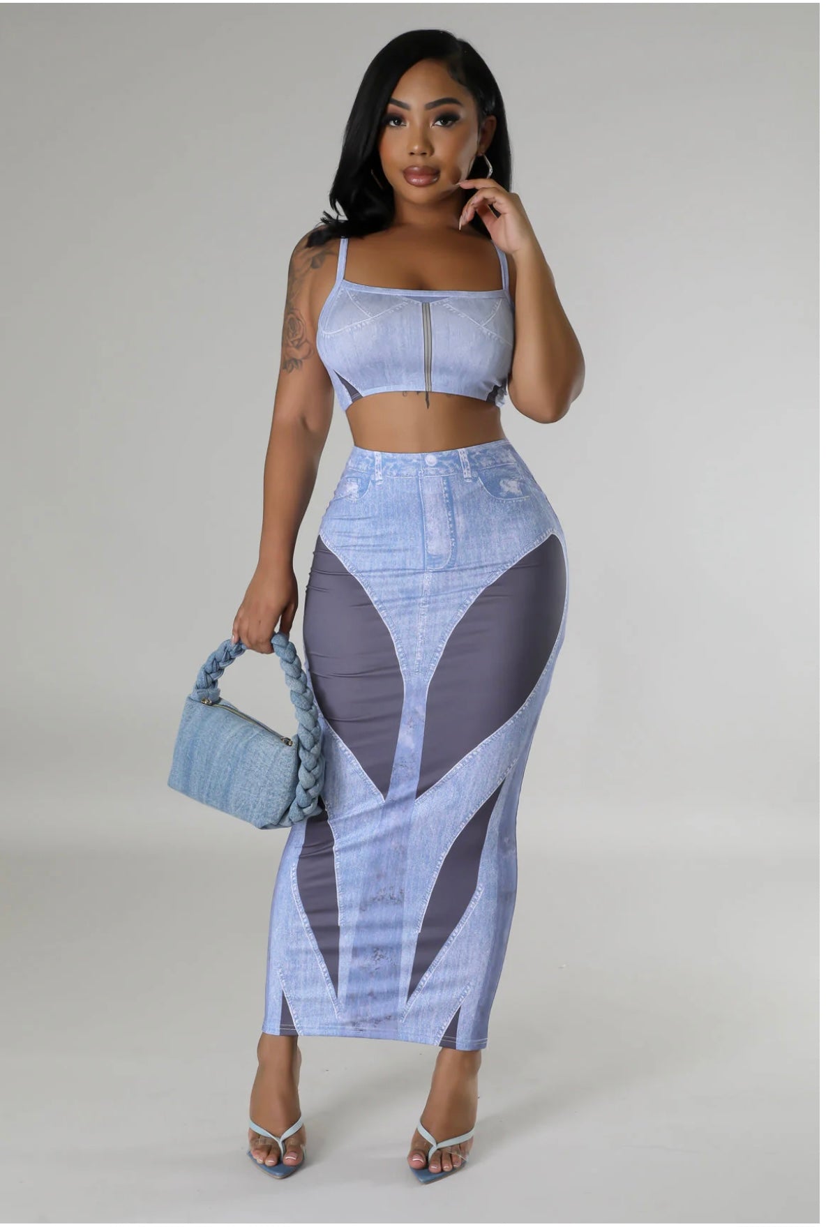 A woman modeling a light blue and gray denim-print two-piece set consisting of a crop top and a high-waisted maxi skirt, holding a matching blue handbag against a neutral gray studio background.